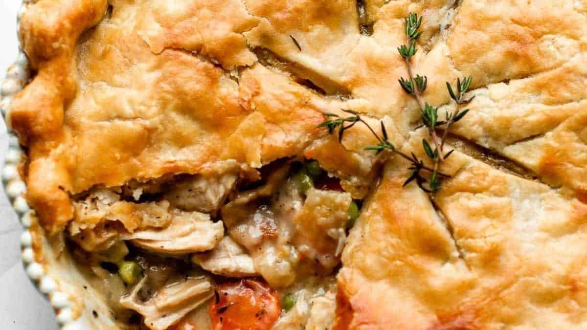 A baked chicken pot pie with a golden-brown crust, one slice removed, showing filling and thyme sprigs on top.