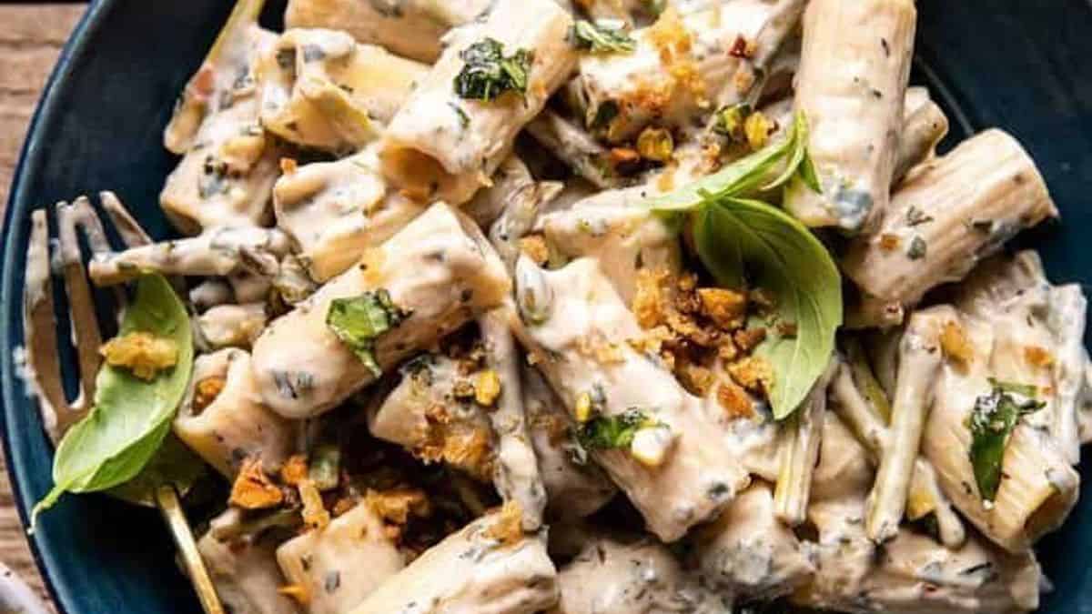 A plate of creamy rigatoni pasta topped with fresh basil leaves and crushed nuts, served with a fork.
