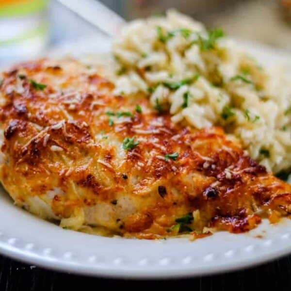 A plate with a baked cheesy chicken breast topped with herbs, served next to a portion of white rice.