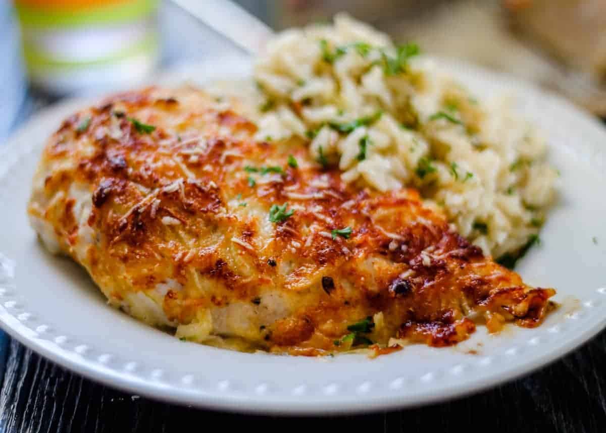A plate with baked, cheese-crusted chicken breast next to creamy rice, both garnished with chopped herbs.