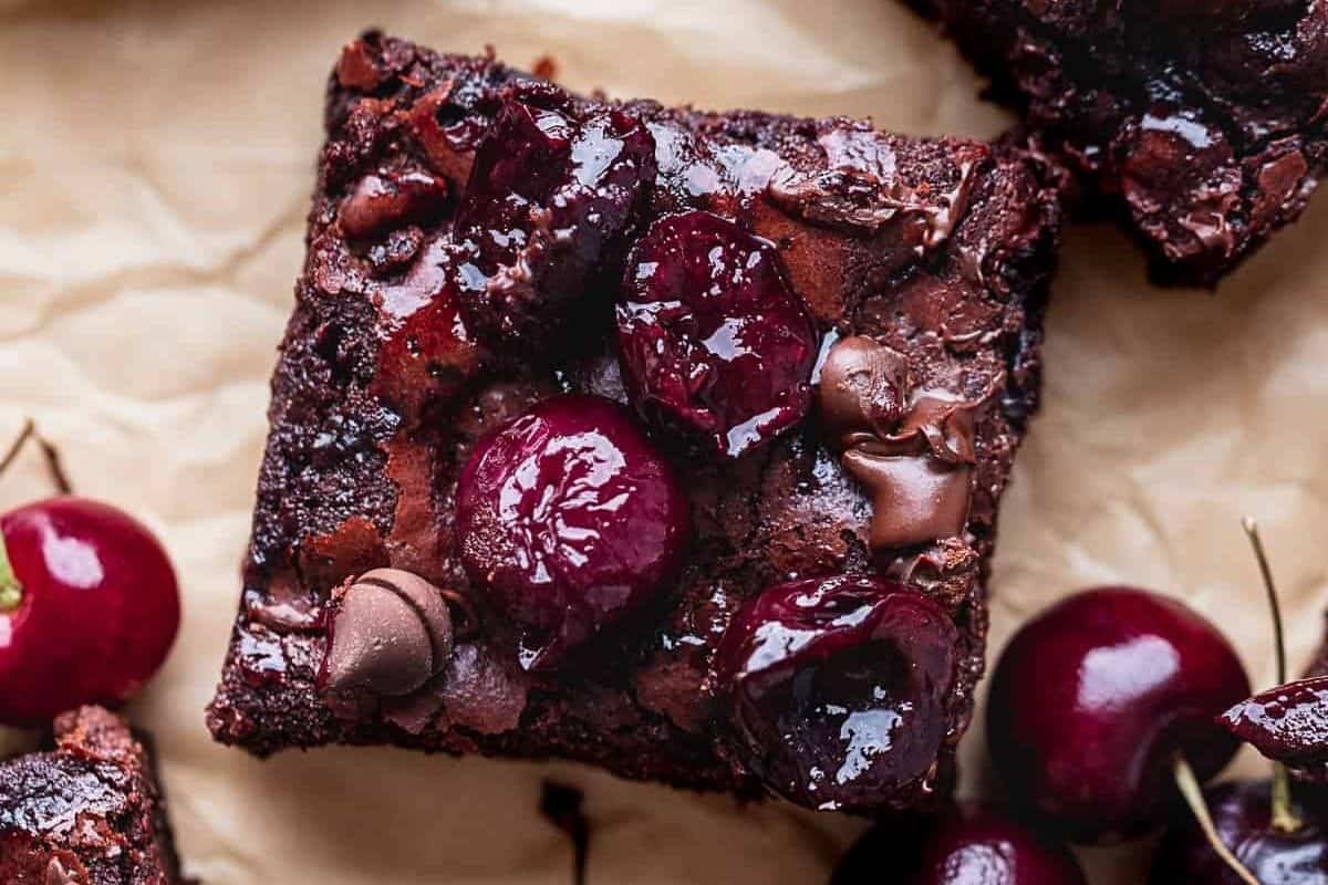 A close-up of a chocolate brownie topped with whole cherries and chocolate chips on crumpled parchment, with fresh cherries nearby.