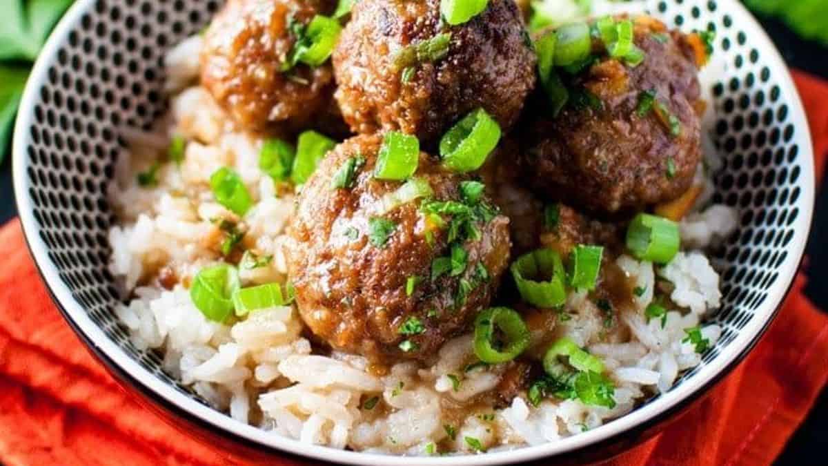 A bowl of white rice topped with meatballs, brown sauce, and garnished with chopped green onions and fresh herbs.