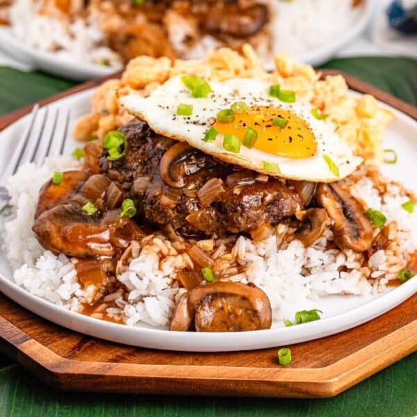 A plate of rice with a hamburger patty, brown mushroom gravy, fried egg, and green onions, served with a fork on a wooden tray.