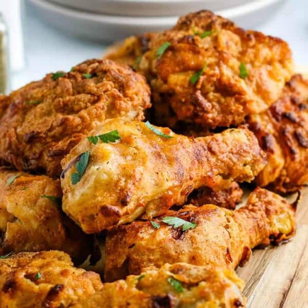 A wooden board with crispy fried chicken garnished with parsley on a marble surface near a jar of seasoning and stacked plates.