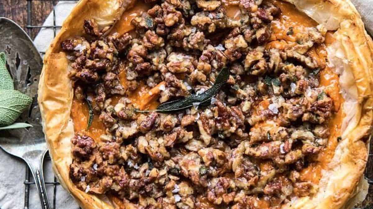 A baked tart with a golden crust, orange filling, topped with chopped pecans and garnished with a fresh sage leaf.