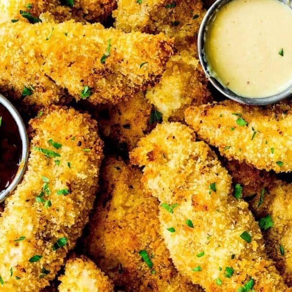 A pile of crispy breaded chicken tenders with two small cups of dipping sauces—one dark, one light—on a parchment-lined plate.