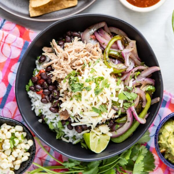 A bowl of rice with shredded meat, black beans, sautéed onions and peppers, cheese, cilantro, lime; sides of corn, guacamole, chips, and salsa.