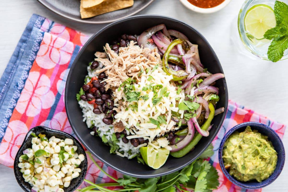 A bowl of rice with shredded meat, black beans, sautéed onions and peppers, cheese, cilantro, lime; sides of corn, guacamole, chips, and salsa.