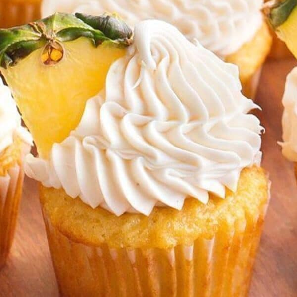 Cupcakes with swirled white frosting and a slice of pineapple on top, arranged on a wooden surface.