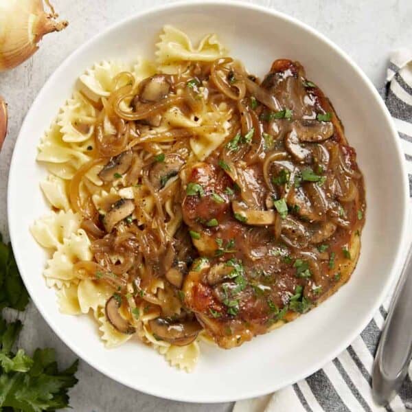 Bowtie pasta with meat, sautéed onions, mushrooms, and brown sauce topped with parsley. Fork, onions, and parsley nearby.