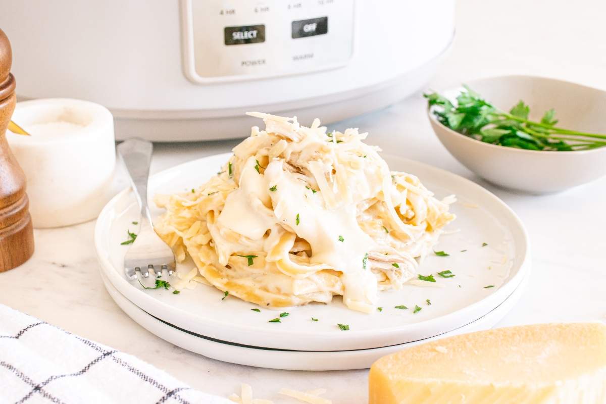 A plate of fettuccine Alfredo with shredded chicken and parsley in front of a slow cooker, with a fork, cheese, and parsley nearby.
