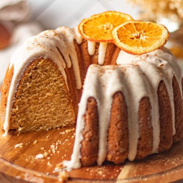 A Bundt cake with white icing drizzled on top, garnished with dried orange slices, sits on a wooden board with a slice removed.