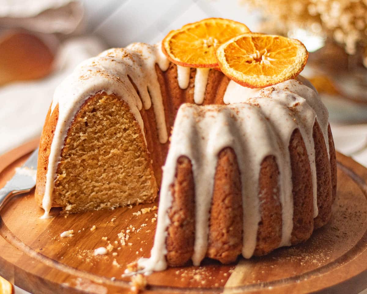 A Bundt cake with white icing drizzled on top, garnished with dried orange slices, sits on a wooden board with a slice removed.