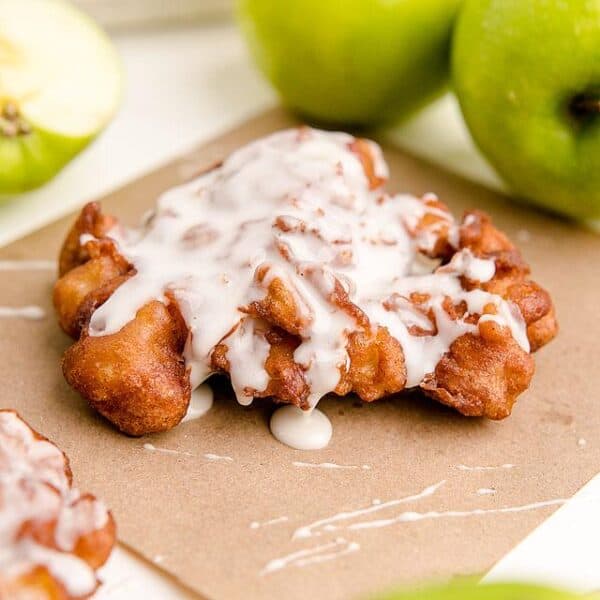 A cinnamon apple fritter with white icing sits on parchment paper, with green apples and a bowl of glaze in the background.