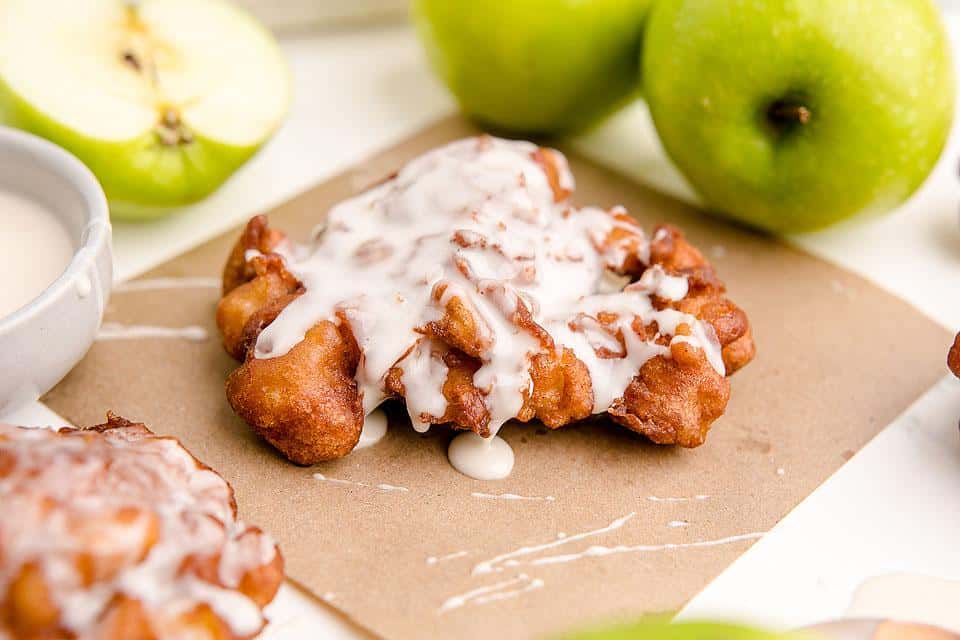 A cinnamon apple fritter with white icing sits on parchment paper, with green apples and a bowl of glaze in the background.