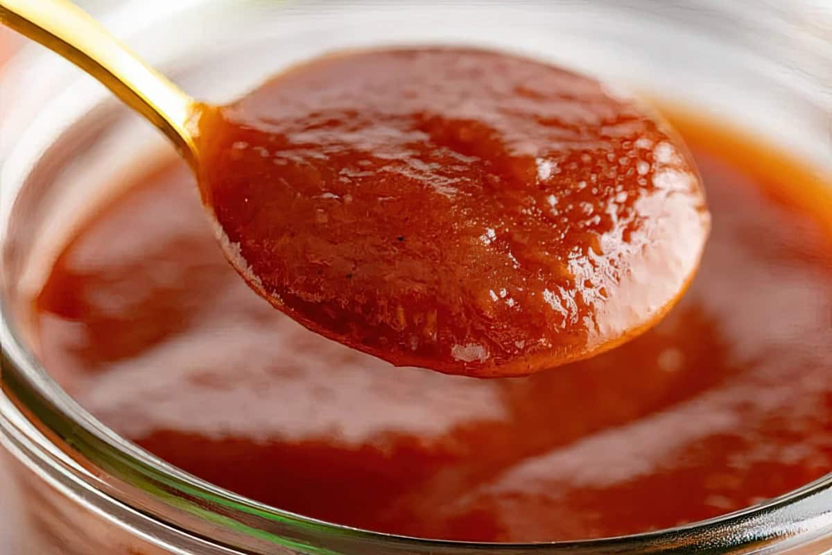 A close-up of a spoonful of thick, red-brown sauce being held above a glass jar filled with the same sauce. The texture appears smooth and glossy, suggesting a rich and flavorful consistency.