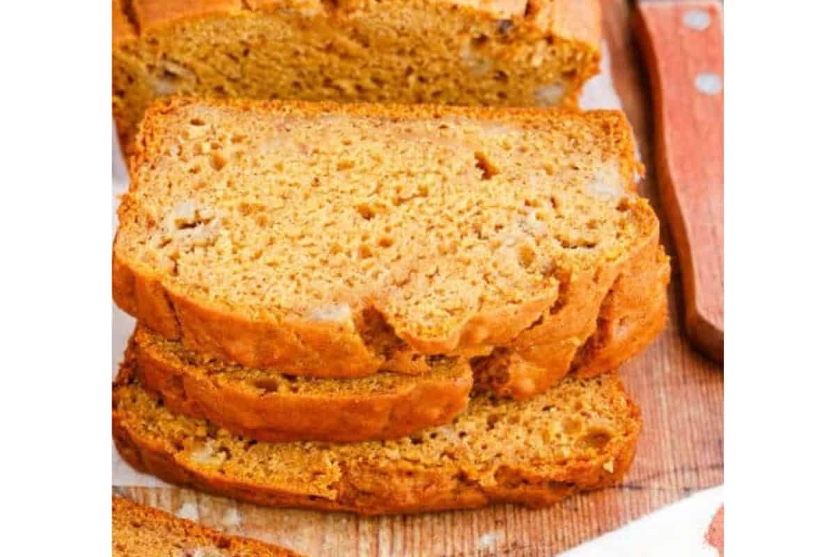 Slices of golden-brown pumpkin bread are stacked on a wooden surface. The bread has a moist and dense texture with small air pockets visible. The warm, cozy appearance of the bread suggests a freshly baked treat, perfect for a fall day.