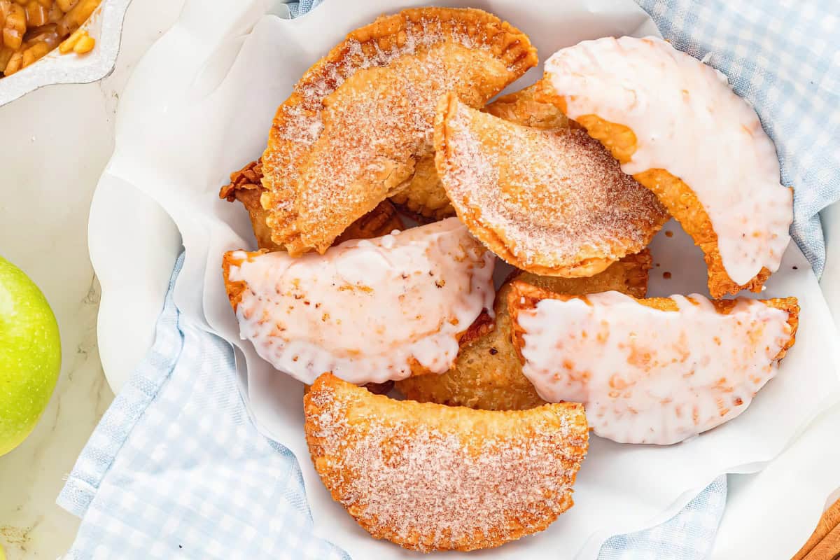 A close-up of several golden-brown, half-moon-shaped fried apple pies topped with a generous sugar glaze, arranged in a white dish lined with a blue-and-white patterned cloth. The background shows a green apple and part of a baking pan.