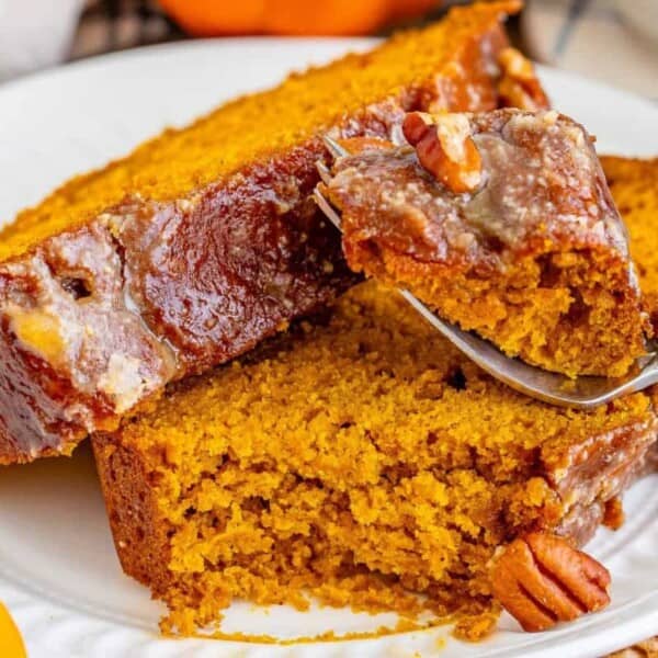 Close-up of a plate with two slices of moist pumpkin bread topped with a pecan glaze. A fork is lifting a bite of the bread, showing its dense, golden crumb and bits of pecan. The plate is set on a wooden table with part of an orange pumpkin visible in the background.