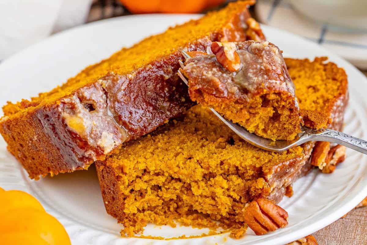 Close-up of a plate with two slices of moist pumpkin bread topped with a pecan glaze. A fork is lifting a bite of the bread, showing its dense, golden crumb and bits of pecan. The plate is set on a wooden table with part of an orange pumpkin visible in the background.