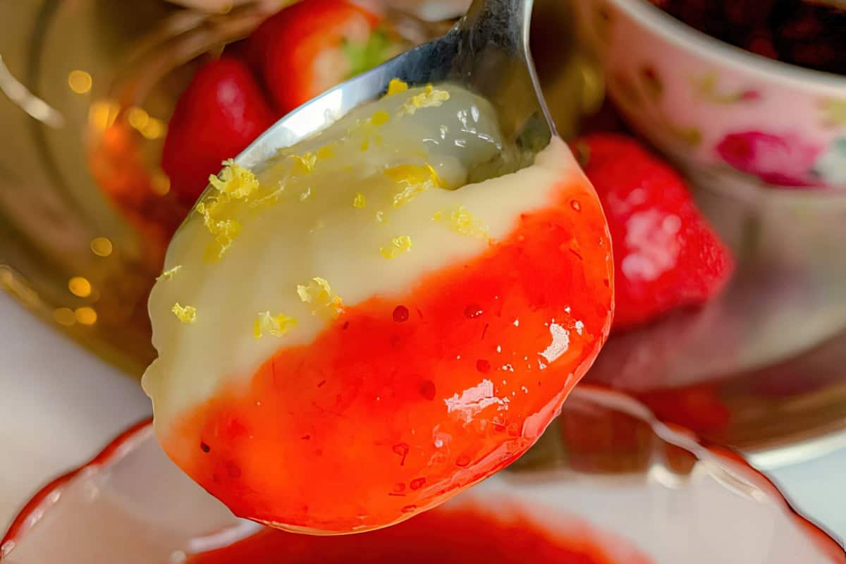 A spoonful of dessert featuring a layer of rich cream topped with a glossy strawberry sauce and lemon zest. In the background, there are fresh strawberries and part of a floral-patterned bowl.