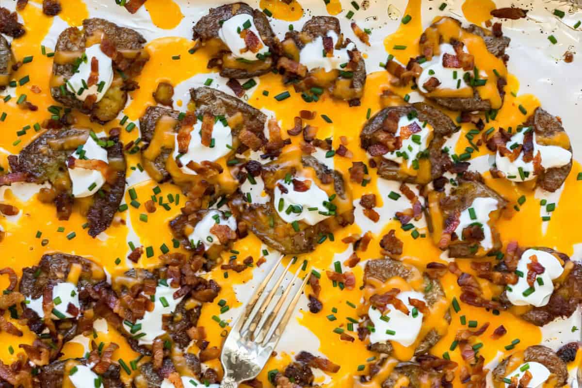A tray filled with loaded potato bites topped with melted cheese, sour cream, chopped chives, and crumbled bacon. A fork rests among the toppings, ready to serve. The golden brown potatoes are evenly spread and covered with the savory toppings.
