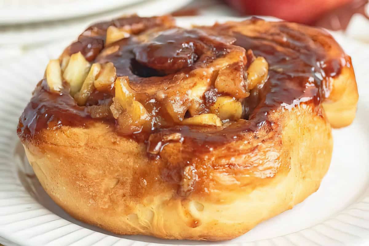 A close-up of a sticky bun on a white plate. The bun is golden-brown and topped with a caramelized glaze and pieces of nuts, making it look rich and sweet.