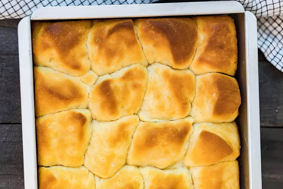 A baking tray filled with golden-brown dinner rolls, each roll closely nestled against the other. The rolls have slightly uneven, fluffy tops with a soft, inviting texture. A checkered cloth is partially visible in the background.