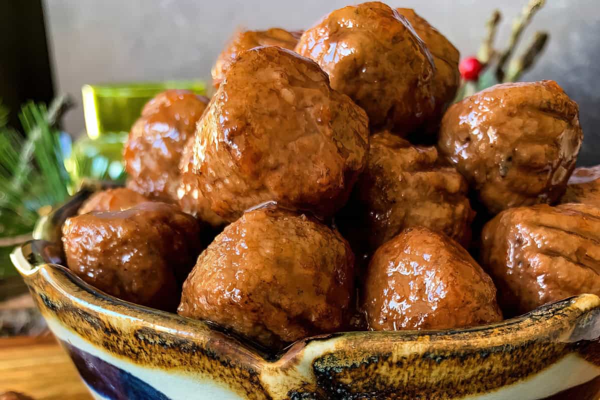 A close-up of a ceramic bowl filled with glazed meatballs. The meatballs are covered in a shiny brown sauce, and the bowl has an earthy tone with a pronounced rim. In the blurred background, there are hints of green foliage and festive decorations.