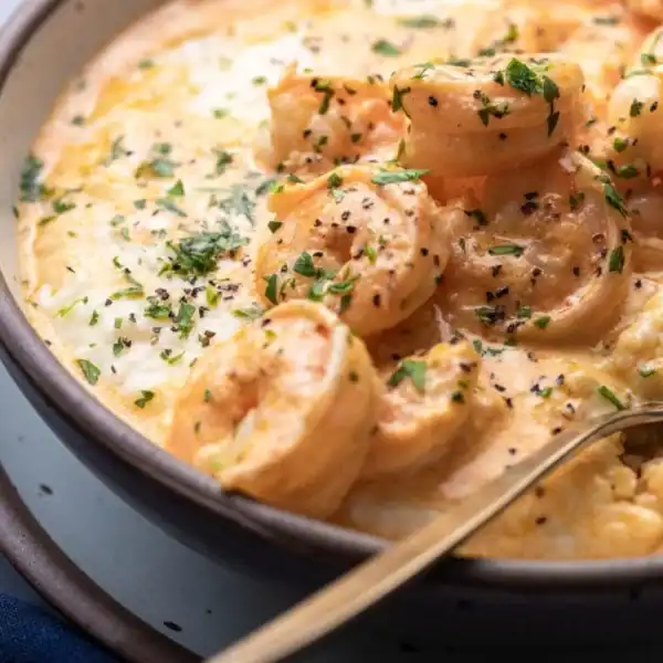 A bowl of shrimp and grits garnished with herbs and black pepper, accompanied by a spoon on the side.