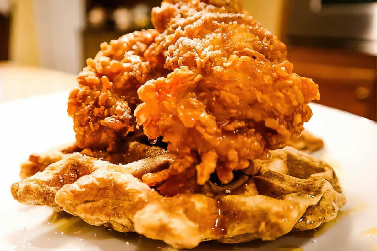 Golden, crispy fried chicken placed on top of a fluffy waffle, with syrup drizzled over the dish, served on a white plate. The background is slightly blurred, highlighting the appetizing meal.