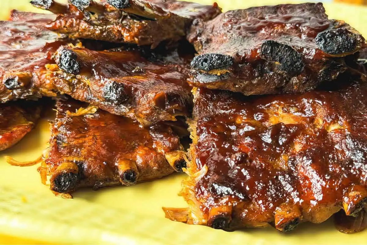 A close-up of a plate filled with saucy, grilled pork ribs, showcasing a deep, glistening barbecue sauce coating. The ribs appear slightly charred and caramelized on the edges, emphasizing their juicy and tender texture. The plate is placed on a yellow surface.