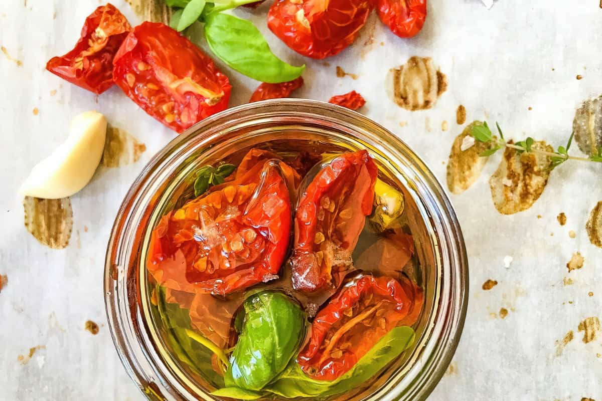 A jar filled with sun-dried tomatoes, basil leaves, and olive oil sits on a parchment paper surface scattered with additional sun-dried tomatoes, garlic cloves, and a sprig of thyme. The tomatoes are vibrant red and partially submerged in the golden oil.
