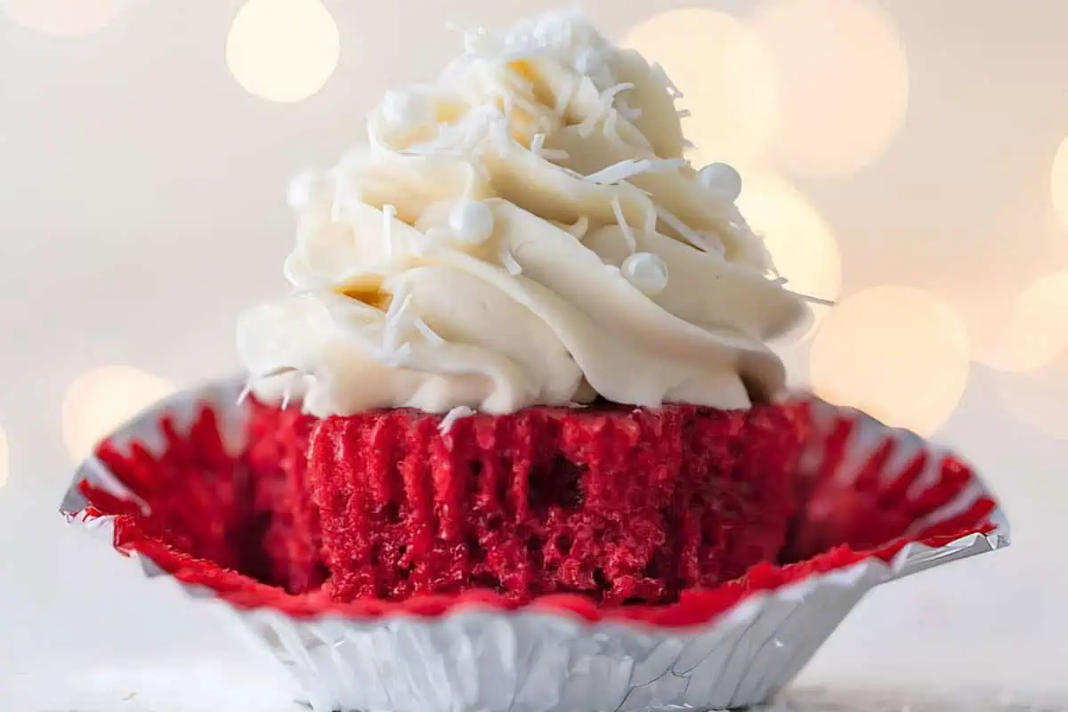 A red velvet cupcake with a bite taken out, revealing its rich, moist interior. The cupcake is adorned with a generous swirl of white cream cheese frosting topped with shredded coconut, all sitting in a partially peeled, red, metallic cupcake liner.