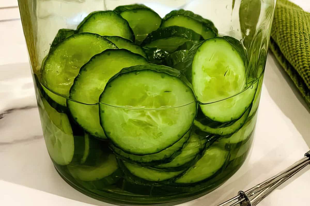 A glass jar filled with rounds of sliced cucumbers. The green cucumber slices are packed tightly together, and the jar sits on a white surface. A green kitchen towel and a metal utensil are partially visible in the background.