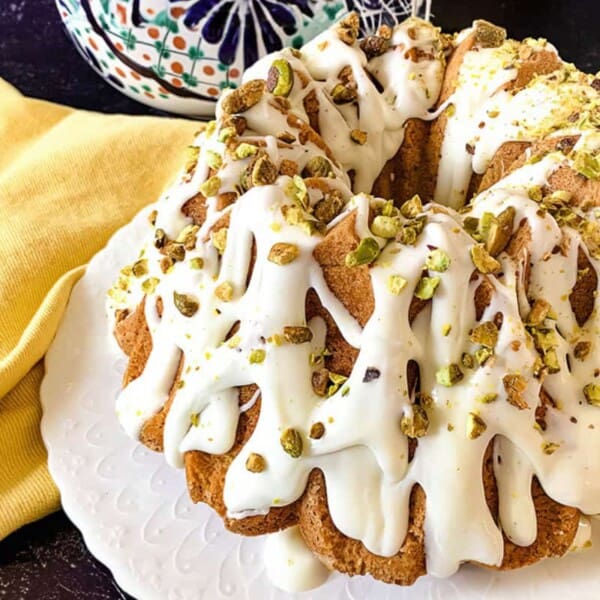 A Bundt cake is topped with white icing and sprinkled with chopped pistachios. The cake is presented on a white plate, with a yellow cloth and a decorative piece of pottery in the background on a dark surface.