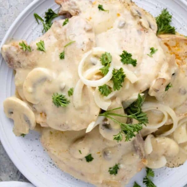 Plate of pork chops with creamy mushroom sauce, onion slices, and fresh parsley on a marble surface next to a striped napkin.