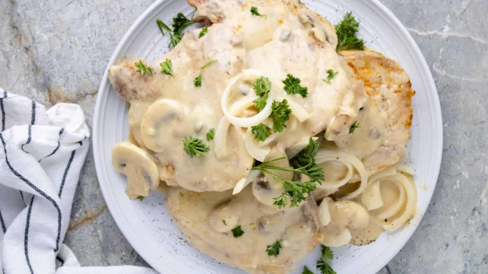 Plate of pork chops with creamy mushroom sauce, onion slices, and fresh parsley on a marble surface next to a striped napkin.