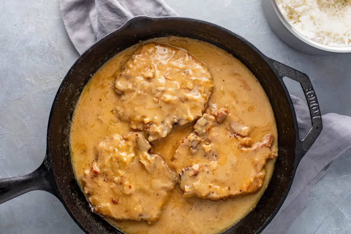 A skillet filled with three pork chops covered in creamy gravy. The skillet is placed on a light grey cloth on a grey surface. In the top right corner, there is a bowl of white rice.