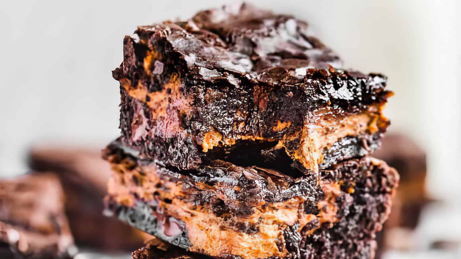 A close-up of two gooey chocolate brownies stacked on top of each other, showing a rich, moist texture with a glossy top layer and swirls of melted chocolate running through the middle. The background is blurred, keeping the focus on the brownies.