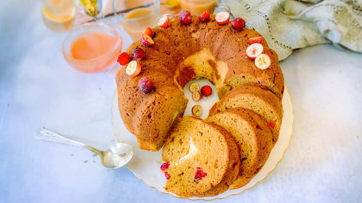 A round bundt cake adorned with small sliced strawberries and whole berries is partially sliced on a white plate. Several pieces are cut and laid out neatly. Three cups with a peach-colored drink and a silver spoon are nearby, along with a light green cloth.