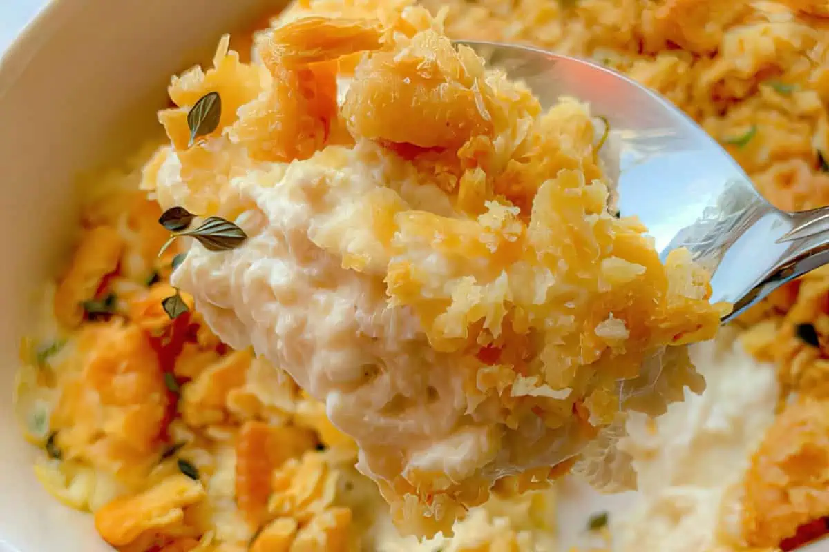 A close-up of a spoonful of creamy macaroni and cheese topped with a golden-brown, crunchy crust. The background shows the rest of the dish in a white bowl, with visible sprigs of fresh thyme for garnish.
