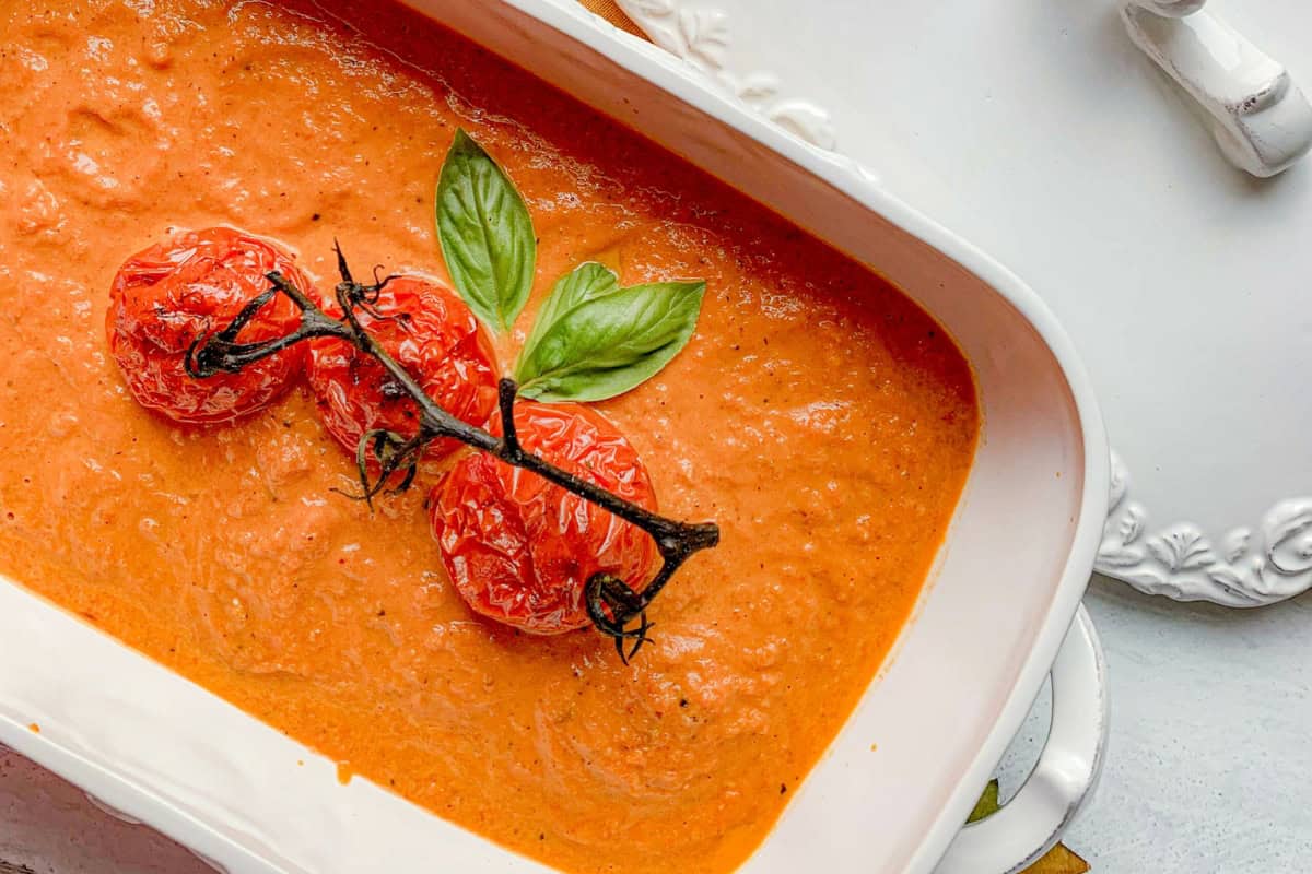 A white dish filled with creamy, orange-red tomato soup garnished with a vine of roasted tomatoes and fresh basil leaves. The dish is placed on a white table next to a lid with a decorative border.