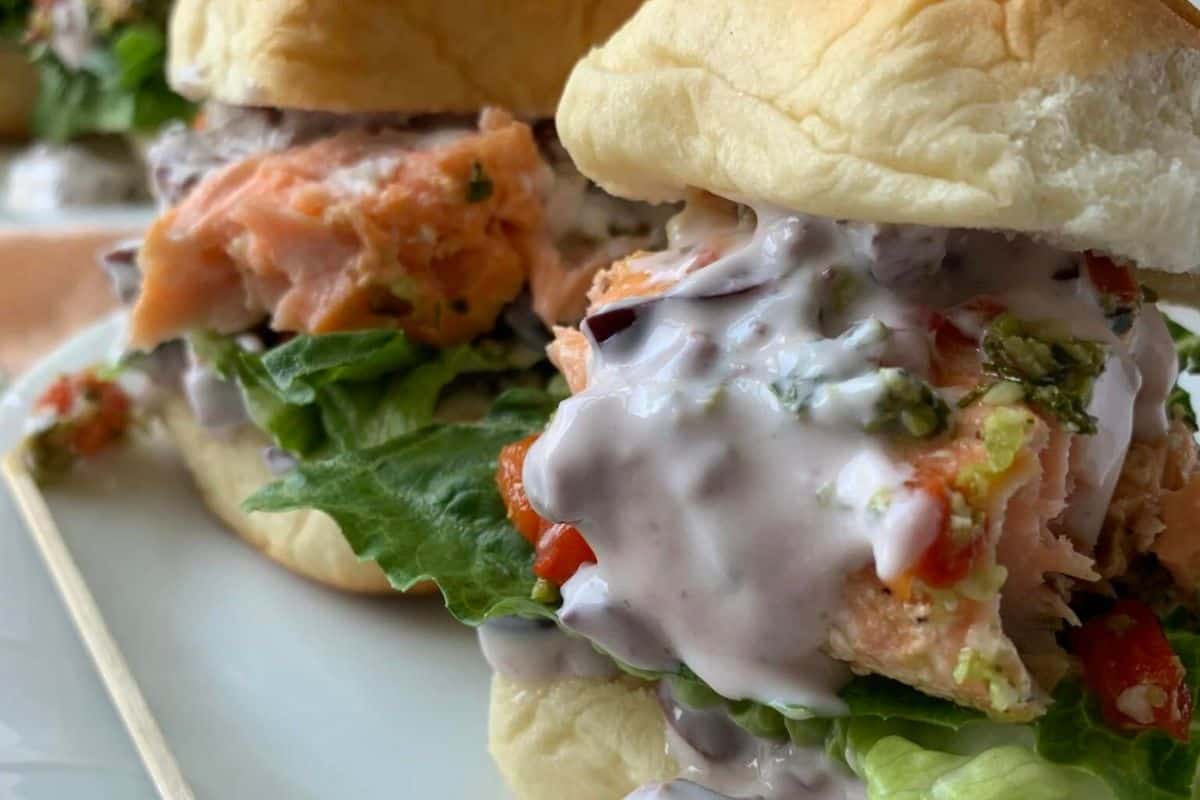 Close-up of two gourmet salmon burgers on soft buns, each filled with fresh lettuce, chunks of salmon, and a creamy purple sauce. The textured composition of the ingredients is highlighted, making the burgers look appetizing and ready to eat.