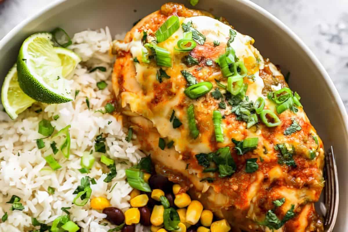 A dish featuring a baked chicken breast topped with melted cheese, green onions, and cilantro, served alongside fluffy white rice with corn and black beans. A lime wedge is also included for garnish. The dish is presented in a gray bowl with a fork.