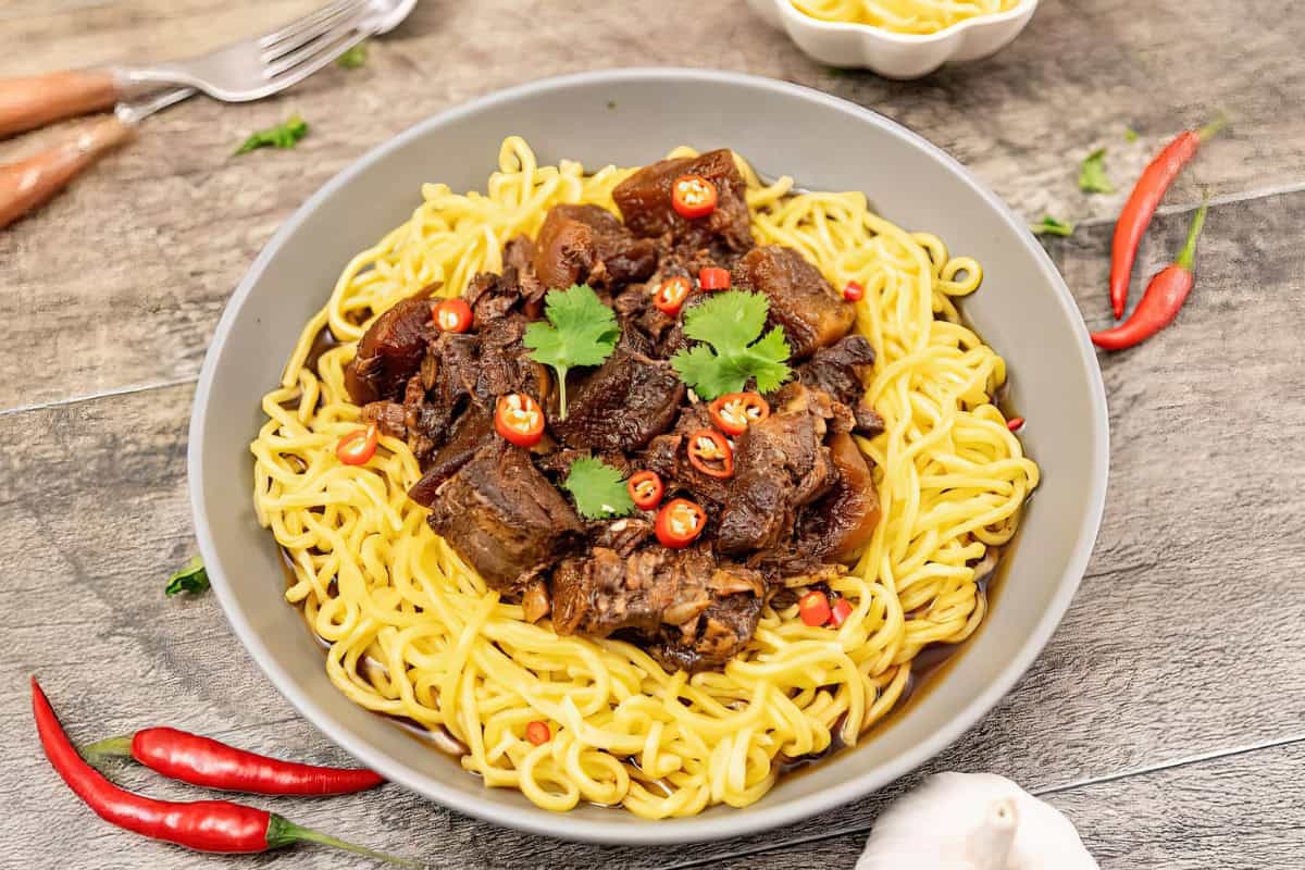 A bowl of yellow noodles topped with stewed beef chunks, garnished with cilantro leaves and red chili slices. The dish is placed on a wooden surface with additional red chilies, a white garlic clove, and a fork and knife visible in the background.