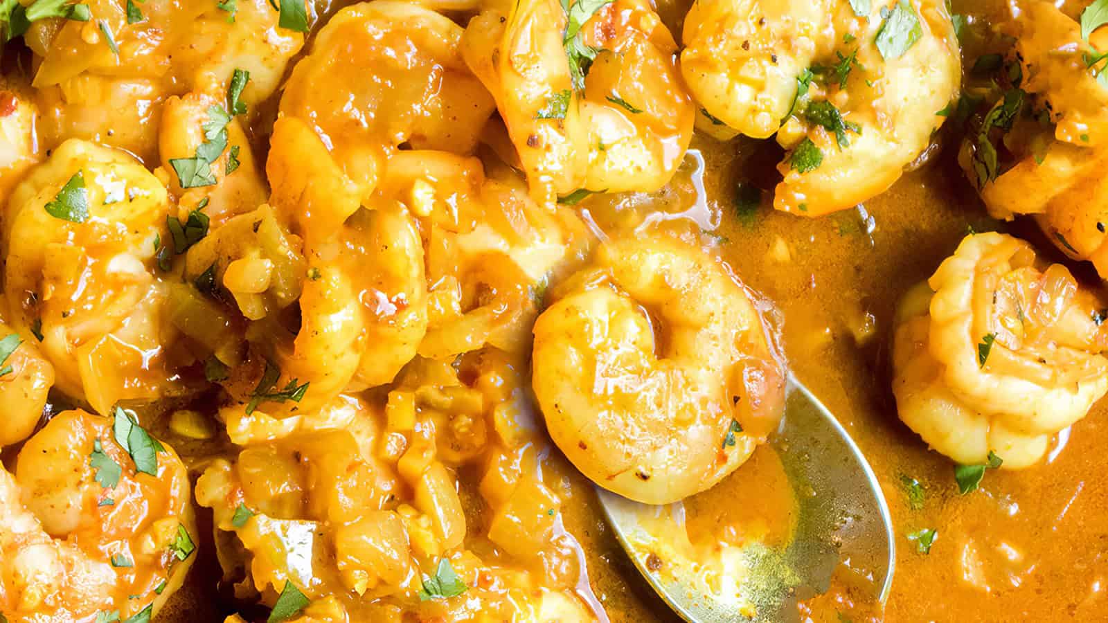 A close-up image of a savory shrimp dish in a rich, orange-colored sauce. The sauce appears to be seasoned with herbs and spices, and there are small garnishes visible. Some shrimp are partially submerged in the sauce, and a spoon is placed on the right side.