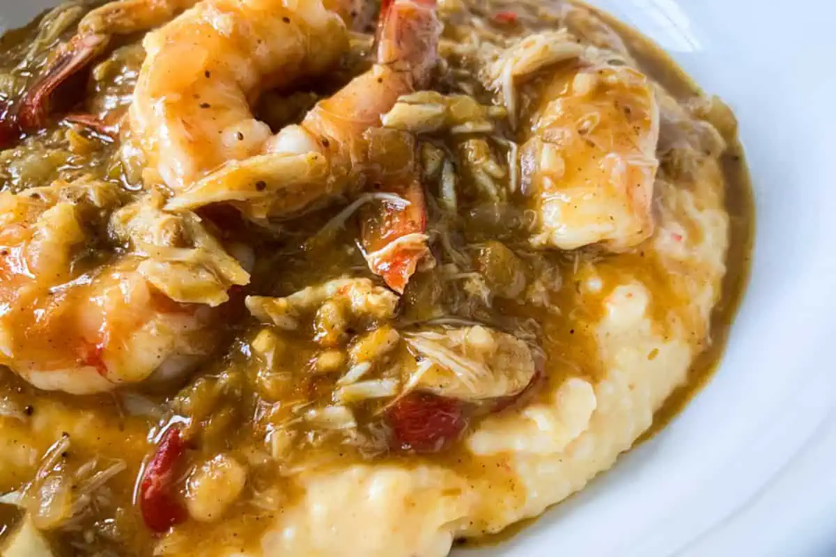 A savory dish of shrimp and grits is shown. Plump shrimp are smothered in a rich, golden gravy with vegetables and possibly crab meat, sitting atop a bed of creamy, buttery grits, creating a hearty, comforting meal.