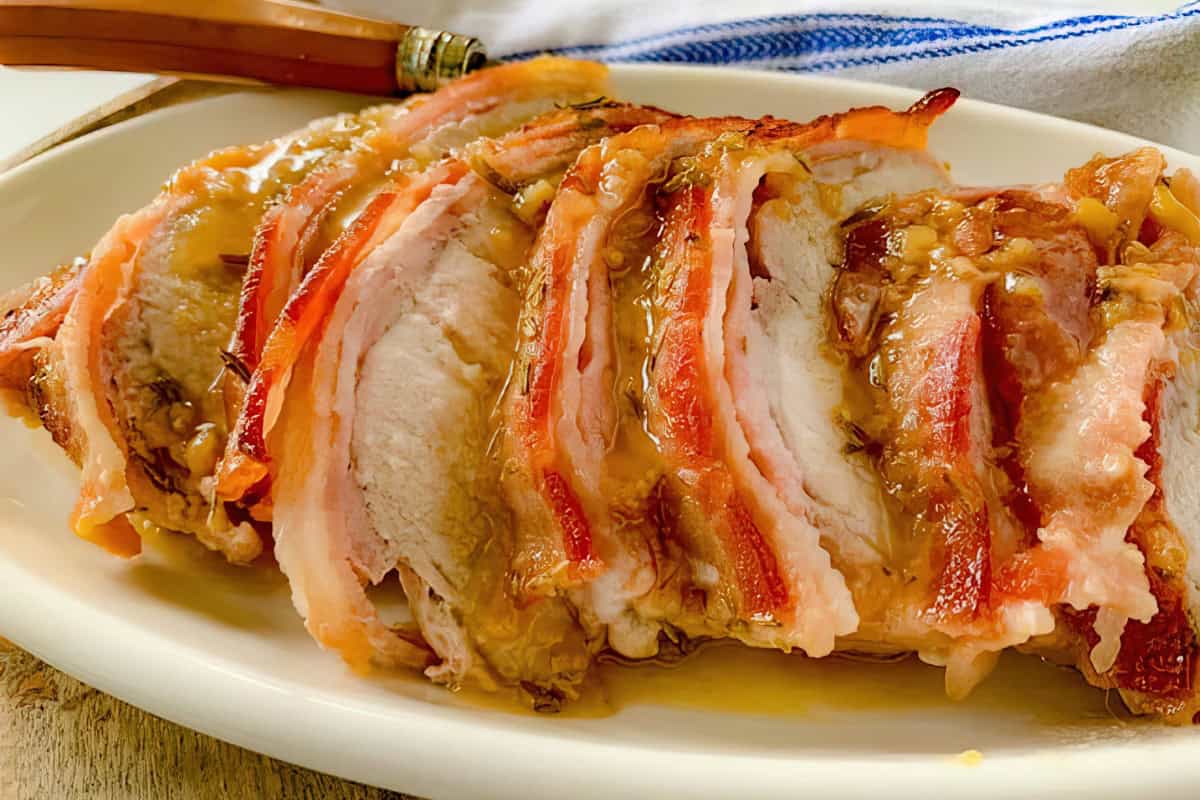 A close-up of a plate of roasted pork tenderloin wrapped in crispy bacon slices and drizzled with a thick sauce. A brown-handled knife is placed on the side of the dish. The pork appears juicy and tender, with a caramelized glaze.