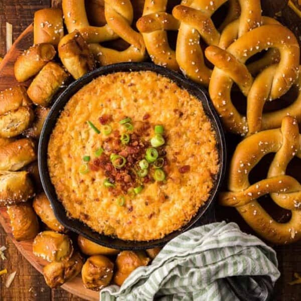 A skillet of cheese dip garnished with bacon bits and green onions, surrounded by soft pretzels, pretzel bites, and grated cheese on a wooden surface.